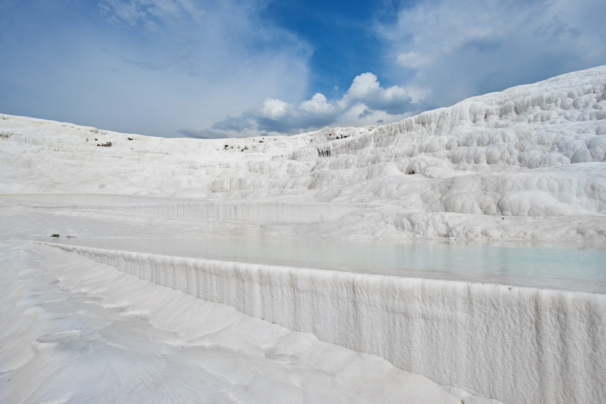 Pamukkale’nin beyaz traverten teraslarında biriken açık mavi durgun su havuzları; parlak beyaz kalsiyum yüzeyleri ve bulutlu mavi gökyüzü ile birlikte geniş bir manzara.